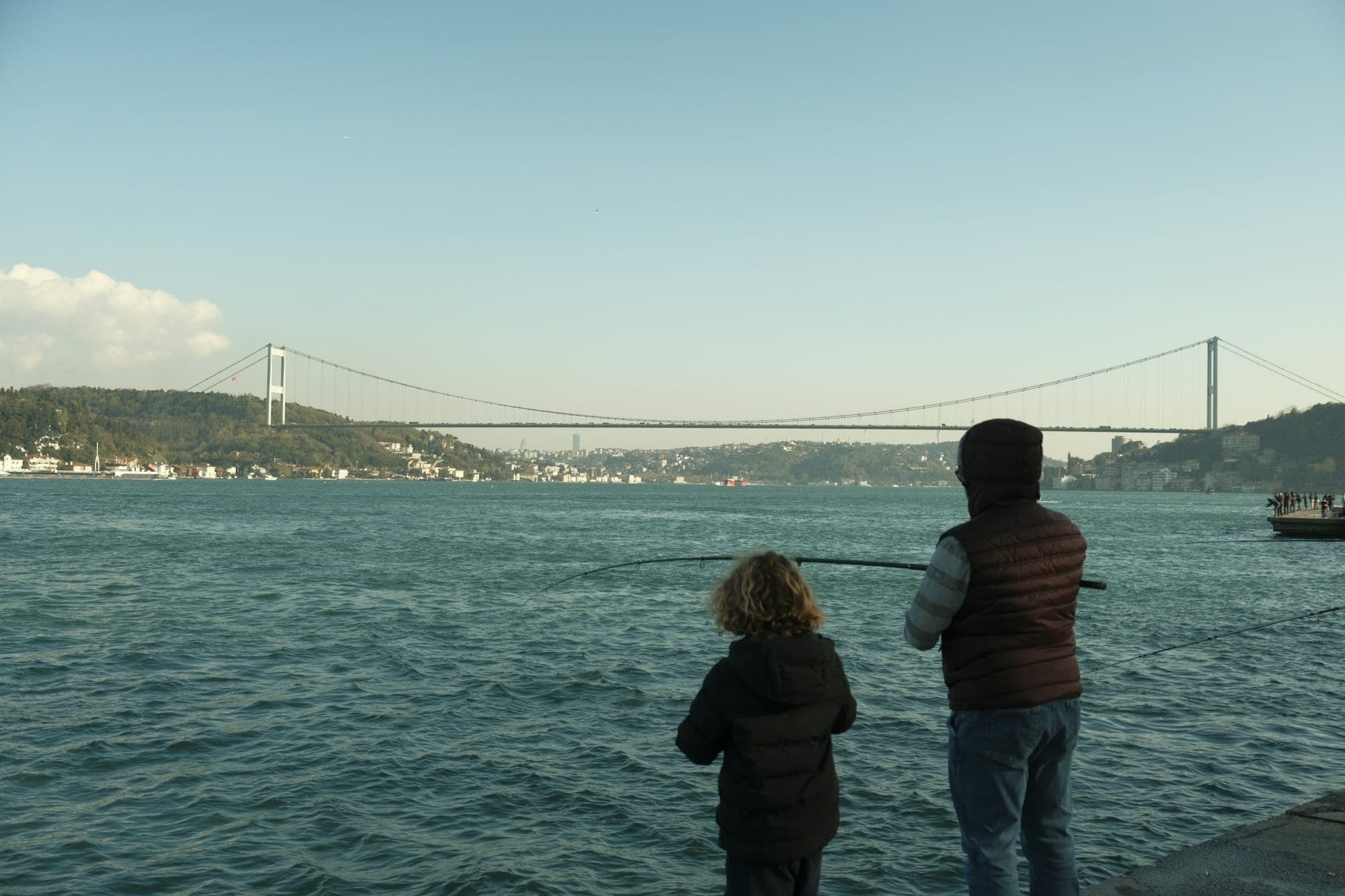 Boğaz kıyısında balık tutan bir baba ve çocuğu, arkada köprü